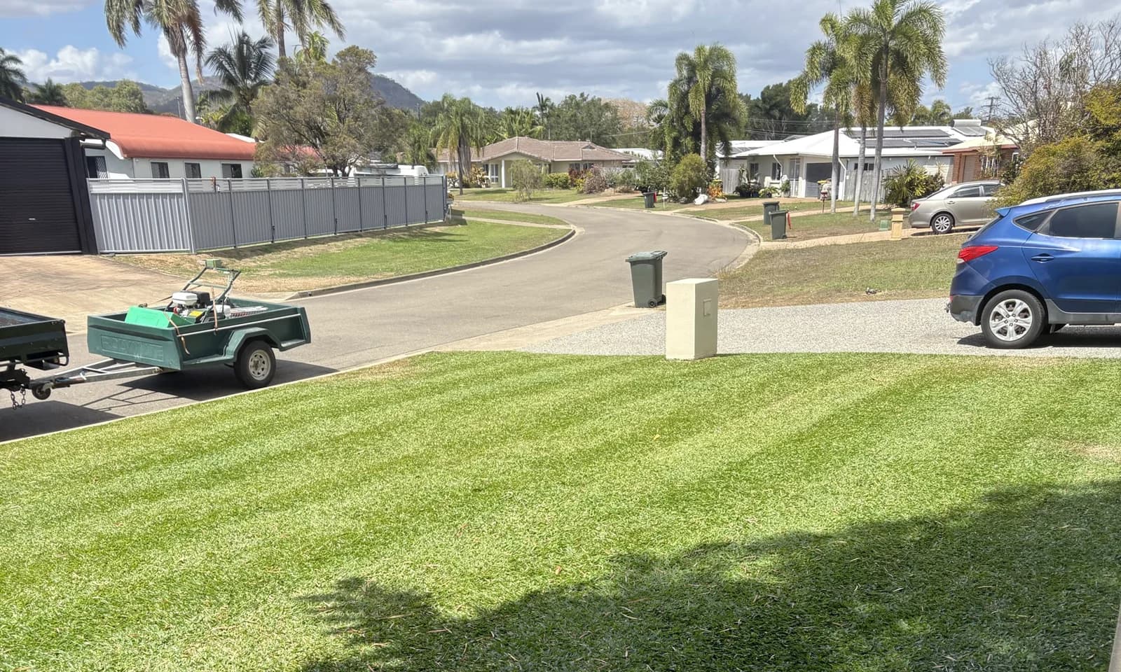 A 6Foot cylinder-mown front lawn in Townsville with fresh stripes across it, the 6Foot trailer and mower on the driveway, palms and the ranges behind